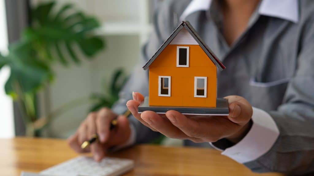 concept of expensive home in current real estate market with high pricing; real estate agent holding out model home in hand